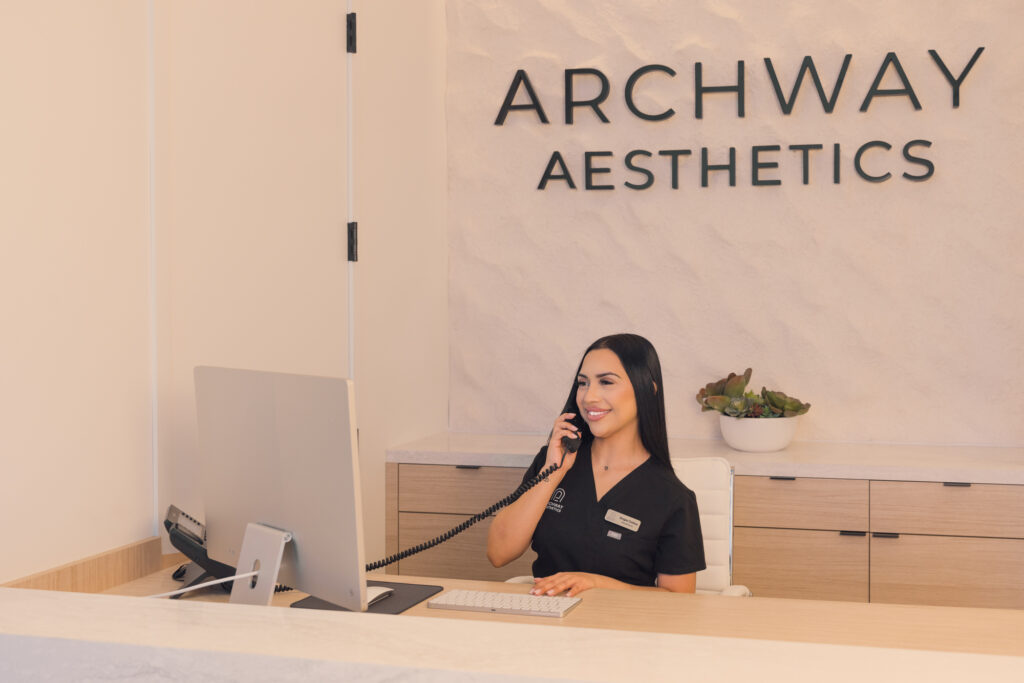A front desk assistant answers the phone at Archway Aesthetics, smiling as she books a Guest for microneedling near Centennial Hills.